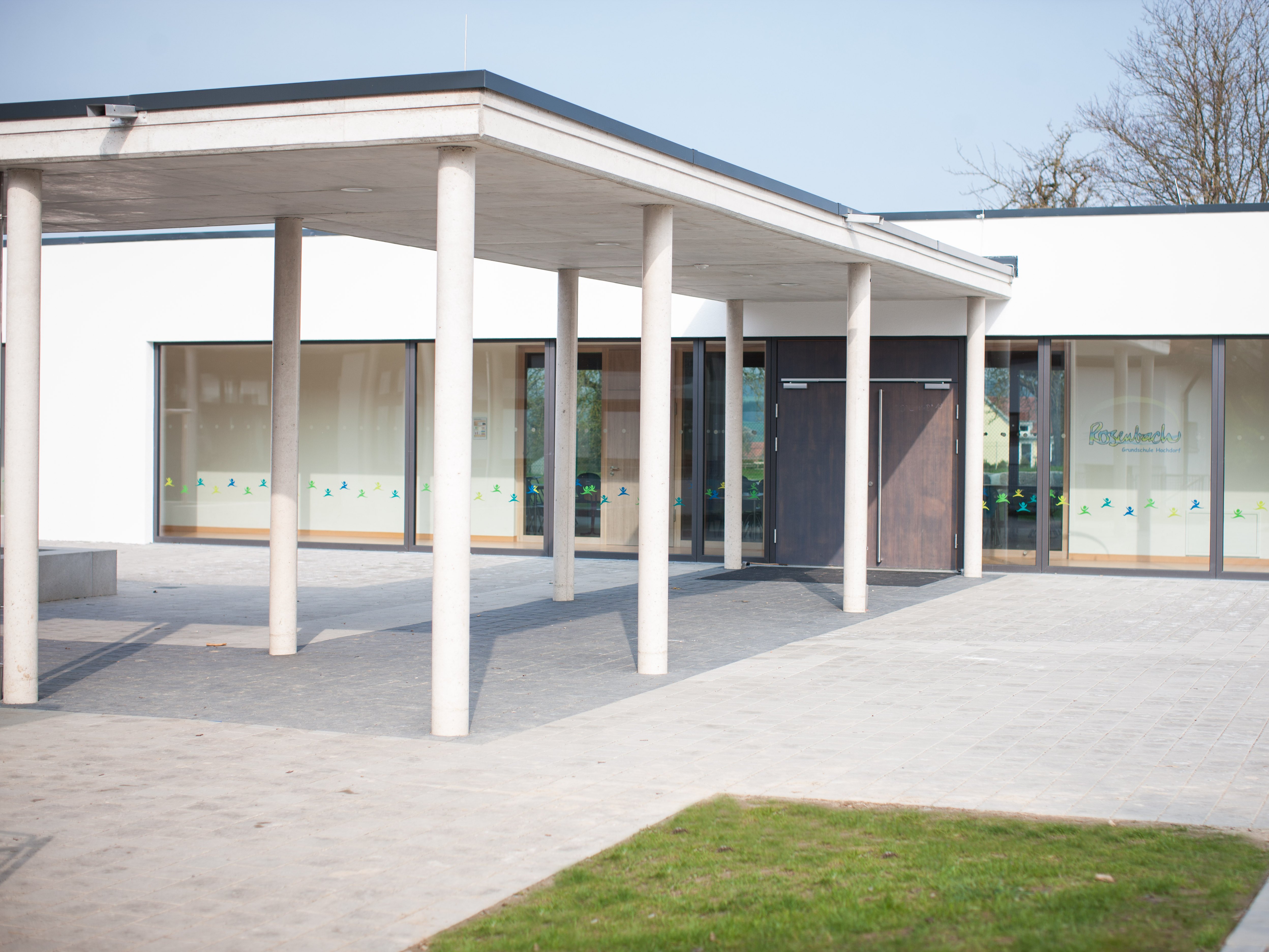 Weiß gepflasterter Pausenhof mit Grünfläche vor dem Eingang eines geradlinigen, neuen Anbaus der Grundschule