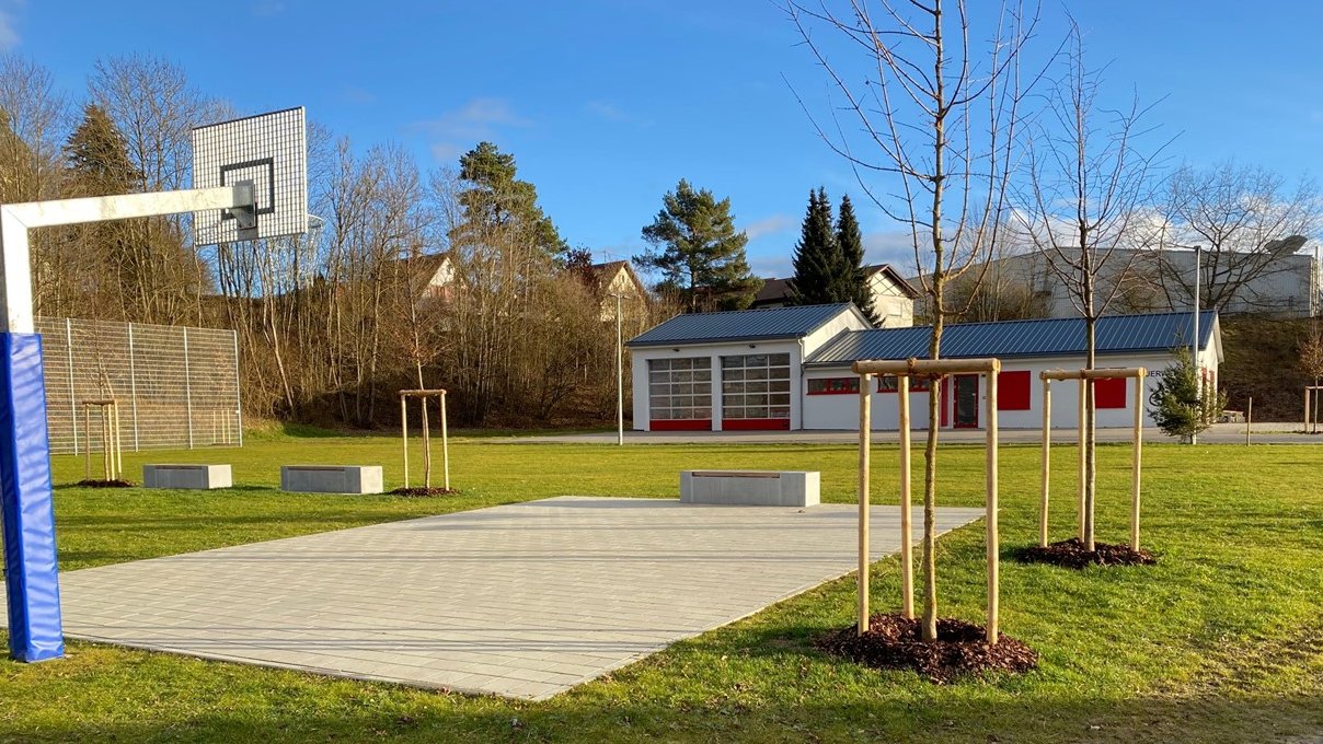 Hartplatz für Basketballspiele umgeben von grünem Rasen und frisch gepflanzten Bäumen im Hintergrund ist ein Kleinspielfeld für Ballspiele und das Feuerwehrhauszu sehen