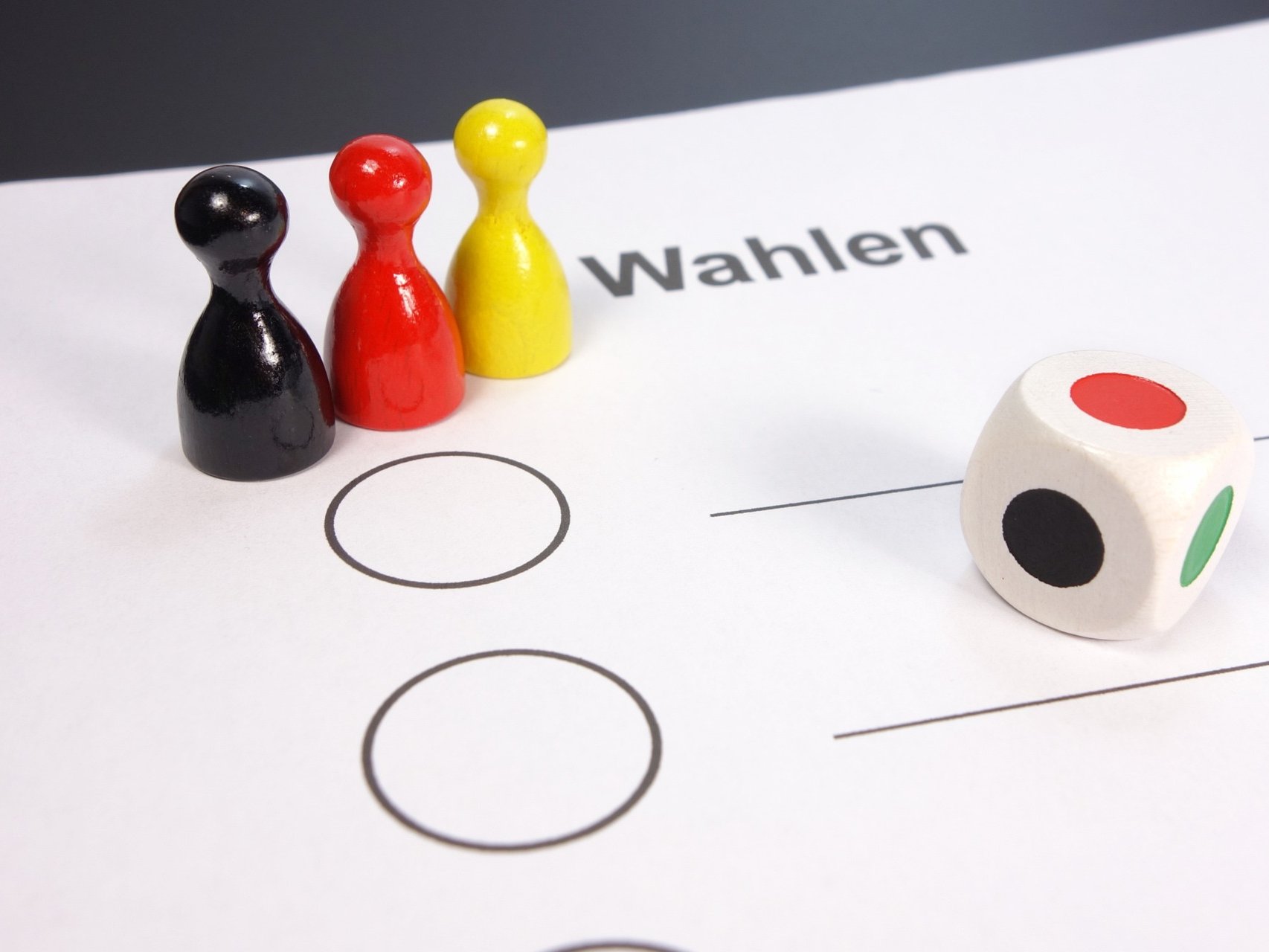 Schwarze, rote und gelbe Spielkegel stehen auf einem Wahlzettel am rechten Rand liegt ein Farbwürfel