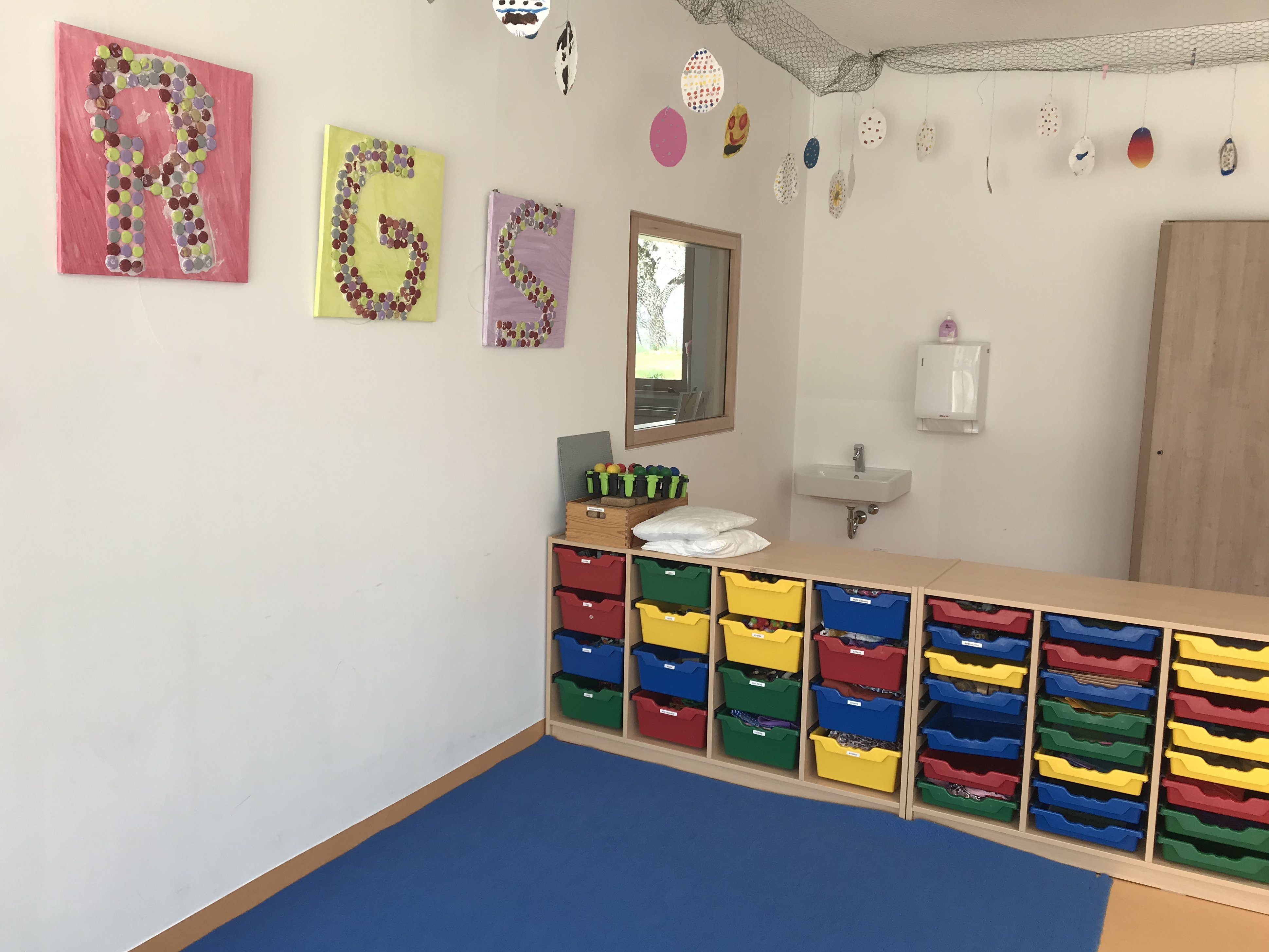 Blick in Betreuungsraum der Rosenbach-Grundschule mit bunte Bilder an weissen Wänden, mit verschiedenfarbigen Ordnungsregalen für Bastelbedarf und blauer Spielmatte auf dem Boden