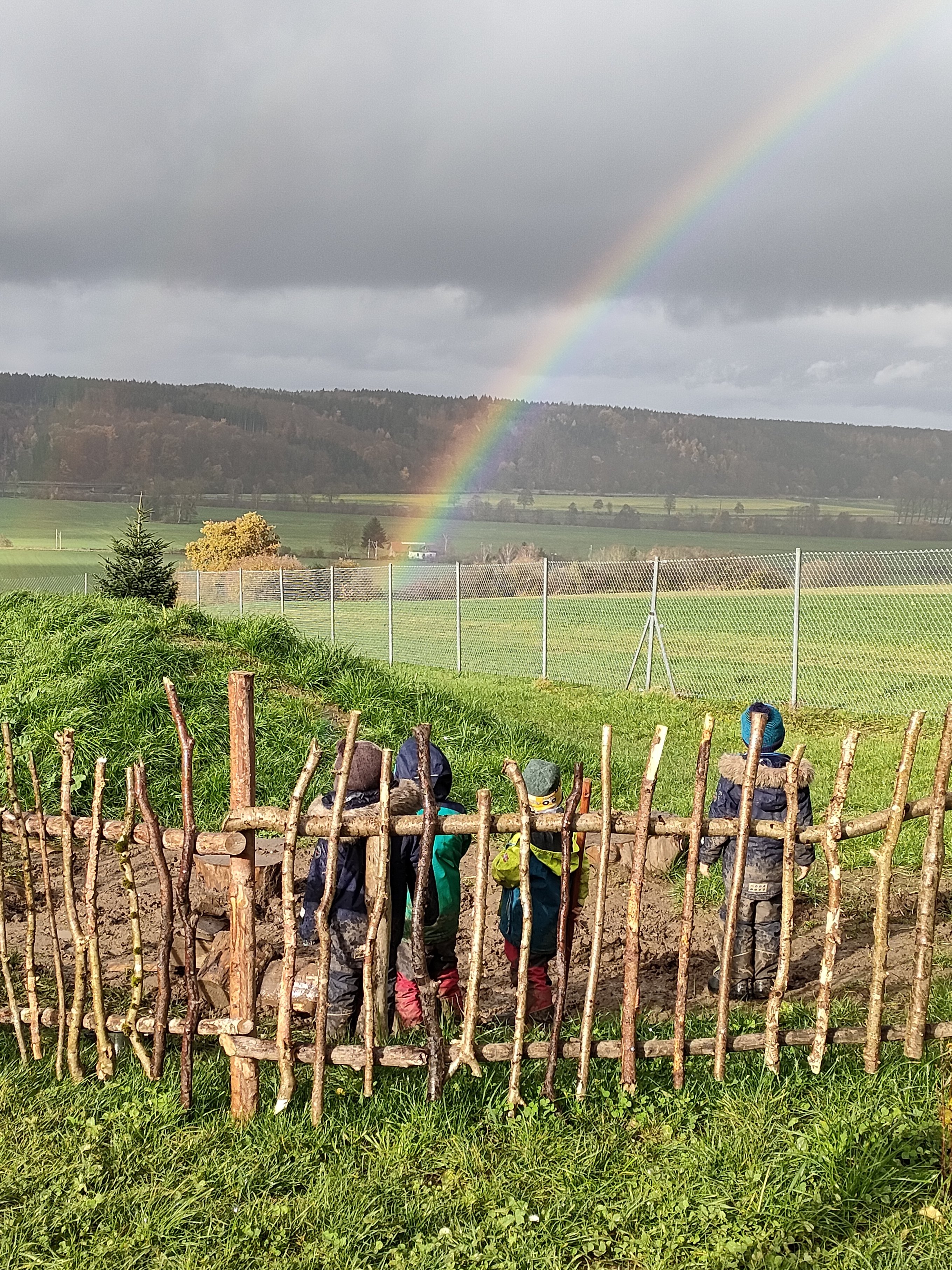 Spielende Kinder auf grüner Wiese bei Nieselregen bewundern einen Regenbogen