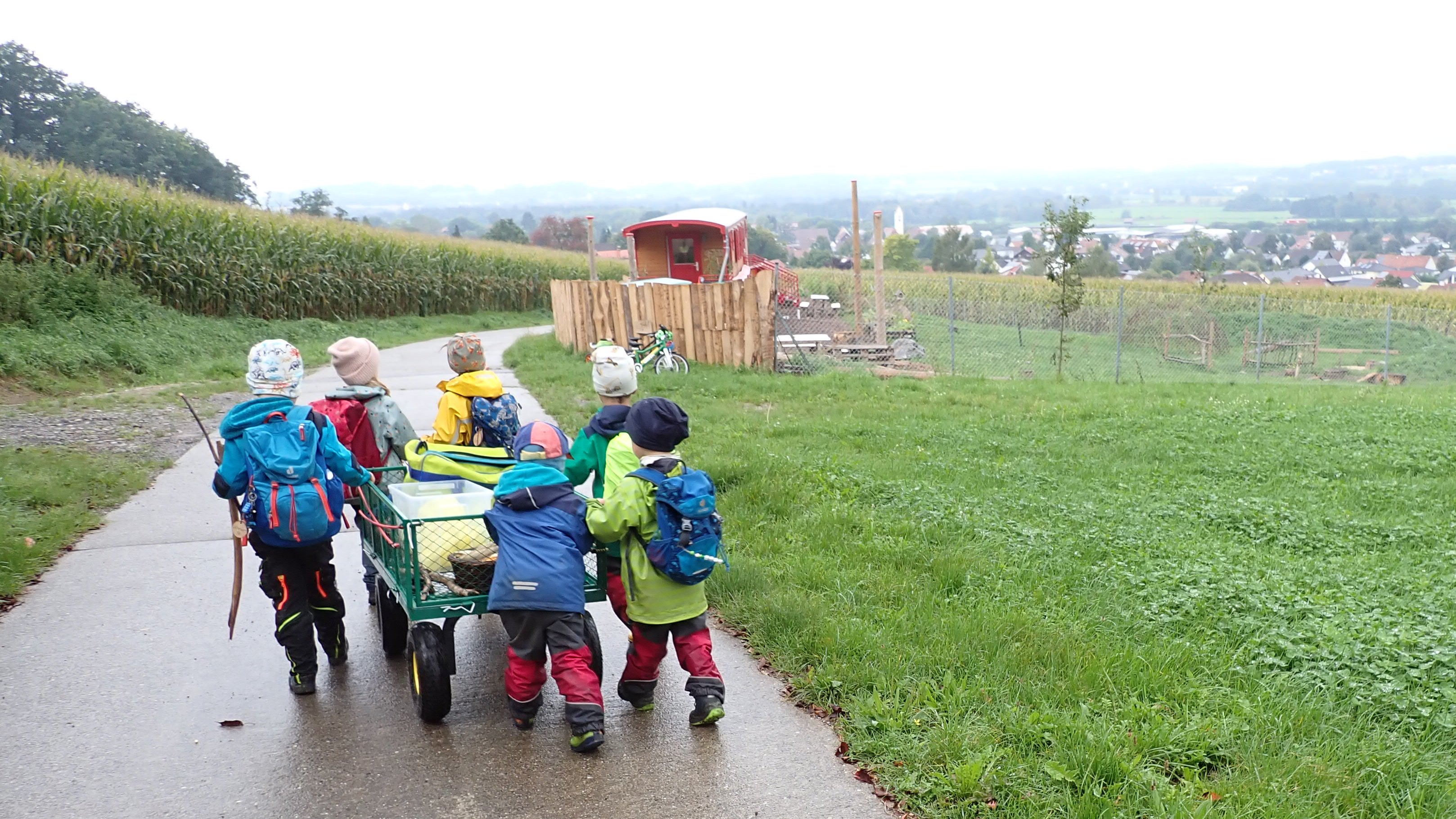 Kinder in bunter Regenbekleidung sind gemeinsam mit einem Bollerwagen im Regen unterwegs