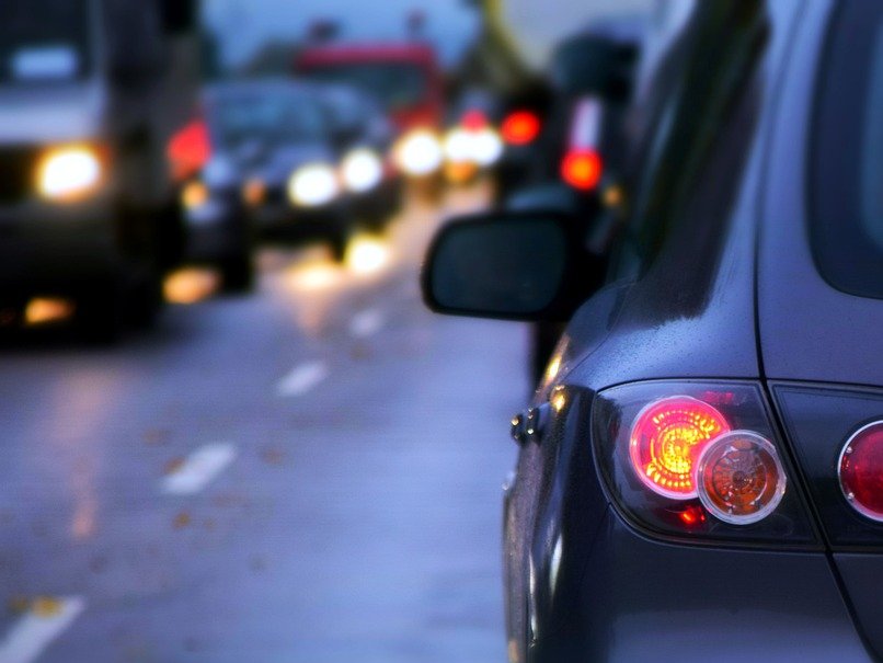 Im Vordergrund ist das rote Rücklicht eines grauen Autos auf einer dunkelblaue Straße zu sehen. Links hinten entgegenkommende Fahrzeuge mit leuchtenden Scheinwerfern zu sehen.
